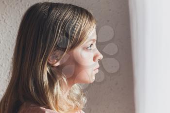 Profile portrait of beautiful blond Caucasian girl looking out the window with white curtains