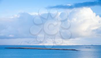 Coastal Icelandic landscape with cargo ship going near small islet and snowy mountains under dramatic blue sky. Reykjavik area, Iceland