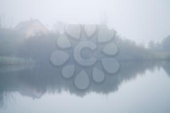 Foggy morning. Small wooden houses on a lake coast, Russian rural landscape