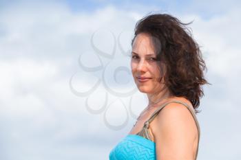 Slightly smiling young adult Caucasian woman in blue dress, outdoor portrait over blue cloudy sky