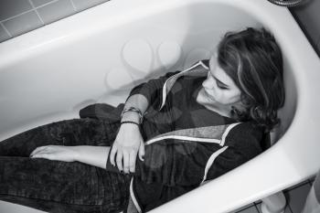 Sad European teenage girl laying in empty shower bath. Depression mood concept, black and white photo