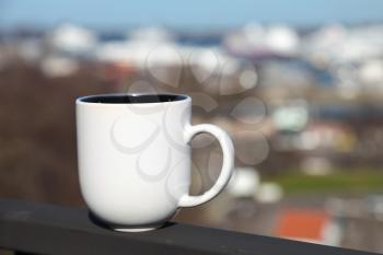 White cup of coffee stands on balcony railing above blurred cityscape