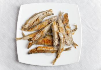 Pile of fried smelts fish lays on a white plate, top view over tablecloth