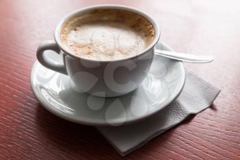 Cappuccino. Cup of coffee with milk foam stands on red wooden table in cafeteria, closeup photo with selective focus 