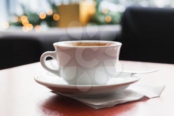 Cappuccino. Cup of coffee with milk foam stands on red wooden table in cafeteria, closeup photo with selective focus and blurred background