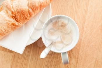 Cappuccino with croissant. Cup of coffee with milk foam stands on wooden table in cafeteria, top view photo with selective focus