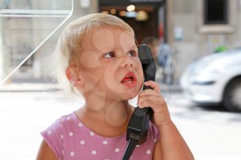 Outdoor portrait of confused little Caucasian blond girl talking on the street phone