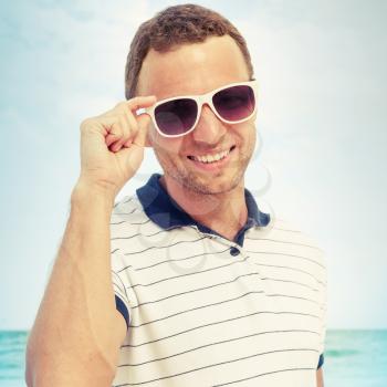Outdoor portrait of young smiling Caucasian man standing with white sunglasses on summer sea coast. Vintage toned square photo with Instagram style photo filter effect