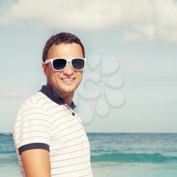 Outdoor portrait of young smiling Caucasian man standing with white sunglasses on summer sea coast. Vintage toned photo with Instagram style photo filter effect