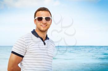 Outdoor portrait of young smiling Caucasian man standing in white sunglasses on summer sea coast