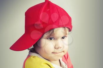 Studio portrait of funny baby girl in red baseball cap over gray wall background. Vintage style, toned photo filter effect