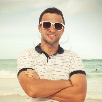 Outdoor portrait of young sporty smiling Caucasian man standing with white sunglasses on summer sea coast. Vintage style toned square photo with filter effect