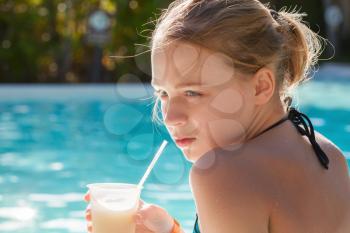 Little blond girl drinks fruit cocktail through plastic tube in swimming pool