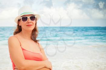 Young happy Caucasian woman in red dress, and sunglasses and white hat. Summer outdoor portrait on the ocean coast. Toned photo with warming filter effect