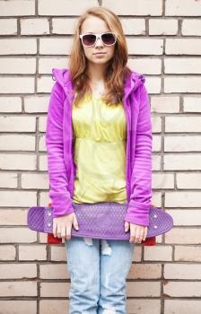 Blond teenage girl in jeans, sunglasses and colorful sporty clothes holds skateboard near by gray urban brick wall