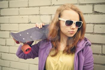 Blond teenage girl in sunglasses holds skateboard near gray urban brick wall, vintage soft tonal correction, old style photo filter effect
