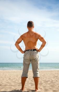 Young sporty sunburned barefoot man dressed in shorts stands on the sandy beach and staring at the sea, back view