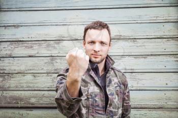 Young Caucasian man in camouflage showing his big fist. Outdoor portrait over green rural wooden wall