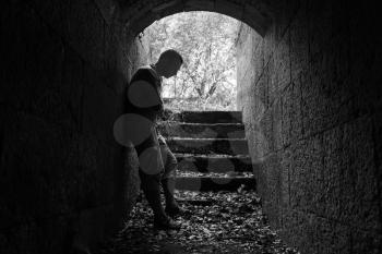 Young sad man stands in dark stone tunnel with glowing end, black and white photo
