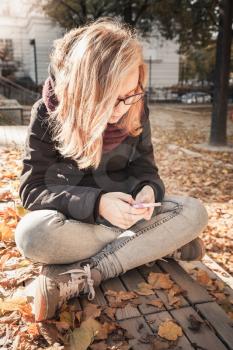 Cute Caucasian blond teenage girl in jeans and black jacket sitting on wooden park bench and using smartphone, outdoor autumn portrait, vintage style tonal correction photo filter