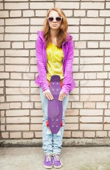 Blond teenage girl in sunglasses and sporty clothes holds skateboard near by gray urban brick wall