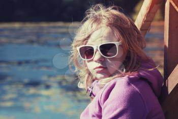 Blond teenage girl in sunglasses sitting on a river coast, outdoor closeup portrait with tonal correction photo filter effect, instagram style