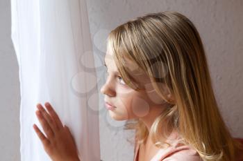 Beautiful blond Caucasian girl looking in a window with white curtains, closeup portrait