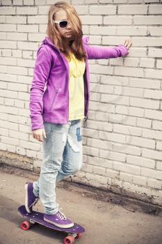 Blond teenage girl in sunglasses and colorful sporty clothes on a skateboard near white urban brick wall. Tonal correction photo filter, old style effect