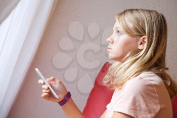 Cute Caucasian blond teenage girl in pink t-shirt using smartphone, indoor closeup profile portrait