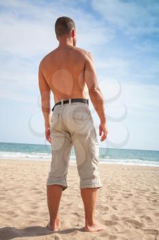 Barefoot sporty man stands on sandy summer beach and looks at the sea