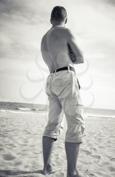 Barefoot sporty man stands on sandy summer beach and looks at the sea, monochrome old style photo