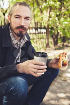 Bearded Asian man having lunch with hot dog and coffee in park, outdoor portrait with selective focus 