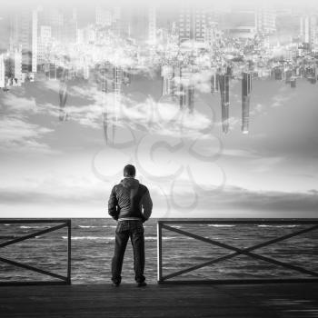 Young man starring on the sea with the city in the sky on a background. Rear view, black and white collage photo