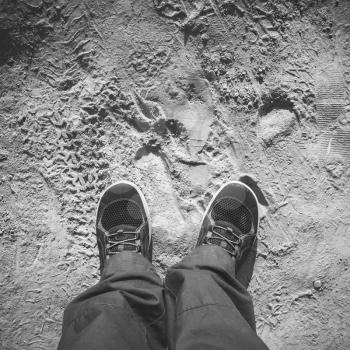 Male feet in sporty shoes stand on dirty dusty road, first person view, square black and white photo