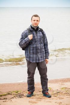 Traveler on Baltic Sea coast. Young adult  Caucasian man in warm outdoor clothes with backpack stands on empty sandy beach