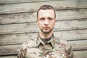 Young serious Caucasian military man in camouflage uniform. Closeup frontal portrait over green wooden wall