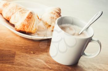 Cappuccino and croissant. White cup of coffee with milk foam stands on wooden table, closeup photo with soft selective focus. Vintage tonal correction filter, old style photo effect