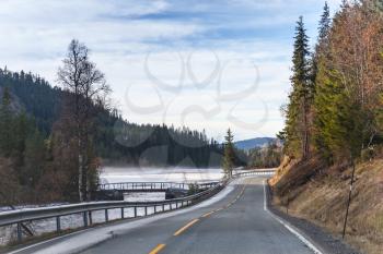 Empty rural Norwegian road goes along frozen mountain river in autumn season