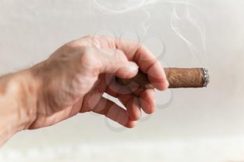 Handmade cigar in male hand, close-up photo with selective focus over white wall background 
