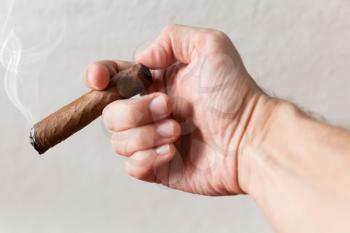 Handmade cigar in male hand, closeup photo with selective focus over white wall background 