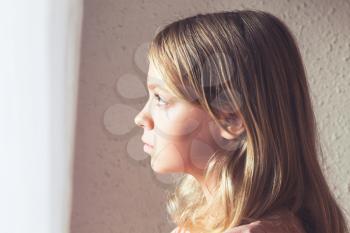 Close-up profile portrait of beautiful blond Caucasian girl looking out the window with white curtains
