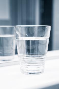 Glass of water stand on white shelf near the mirror, blue toned vertical close-up photo
