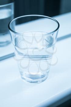 Glass of water stand on shelf near the mirror, blue toned vertical closeup photo