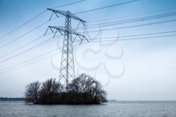 Transmission power tower, electricity pylon stands on small island with bare trees. Steel lattice tower, used to support an overhead power line