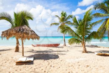 Palm trees, empty loungers, boat and umbrella are on white sandy beach. Caribbean Sea, Dominican republic, Saona island coast, touristic resort