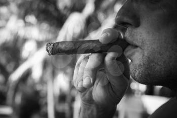 Young European man smokes big cigar, close up black and white photo with selective focus. Dominican Republic