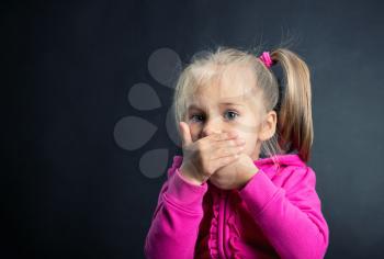 Little girl covers her mouth with hands on dark background