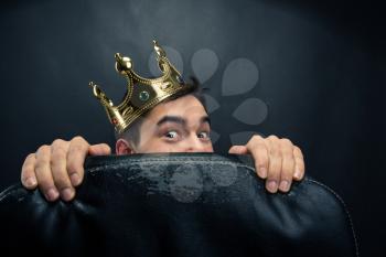 Scared man with crown on the head hidding behind the chair