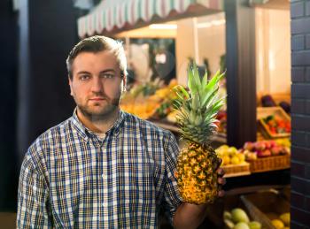 Portrait of smiling man in shirt holds ripe pineapple in hands. Grocery on the background.