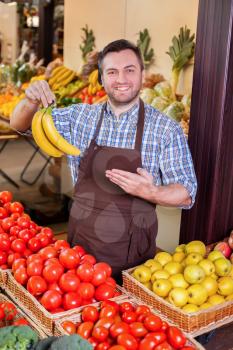 Smiling man offers fresh bananas in in front of boxes with tomatoes and yellow apples. Grocery on the background.